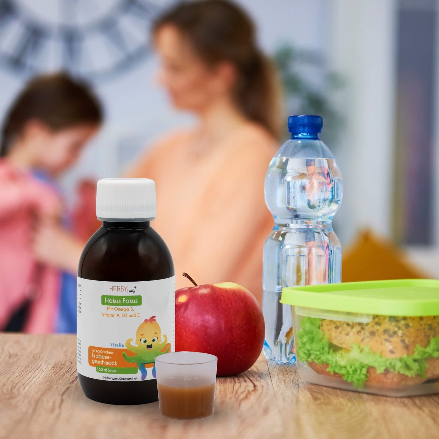 Hokus Fokus Sirup mit Dosierbecher steht neben einem Apfel, einer Wasserflasche und einer Brotdose, im Hintergrund hilft eine Mutter ihrer Tochter beim Jacke anziehen