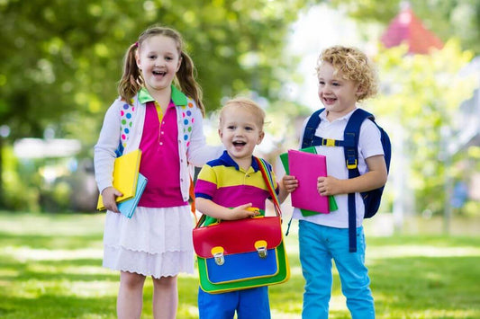3 Kinder stehen auf Wiese mit Bäumen im Hintergrund. Alle tragen einen bunten Rucksack oder Schultasche 